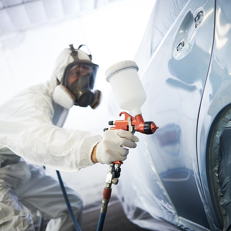 Carrossier en combinaison et masque à gaz appliquant de la peinture sur une voiture avec un pistolet.