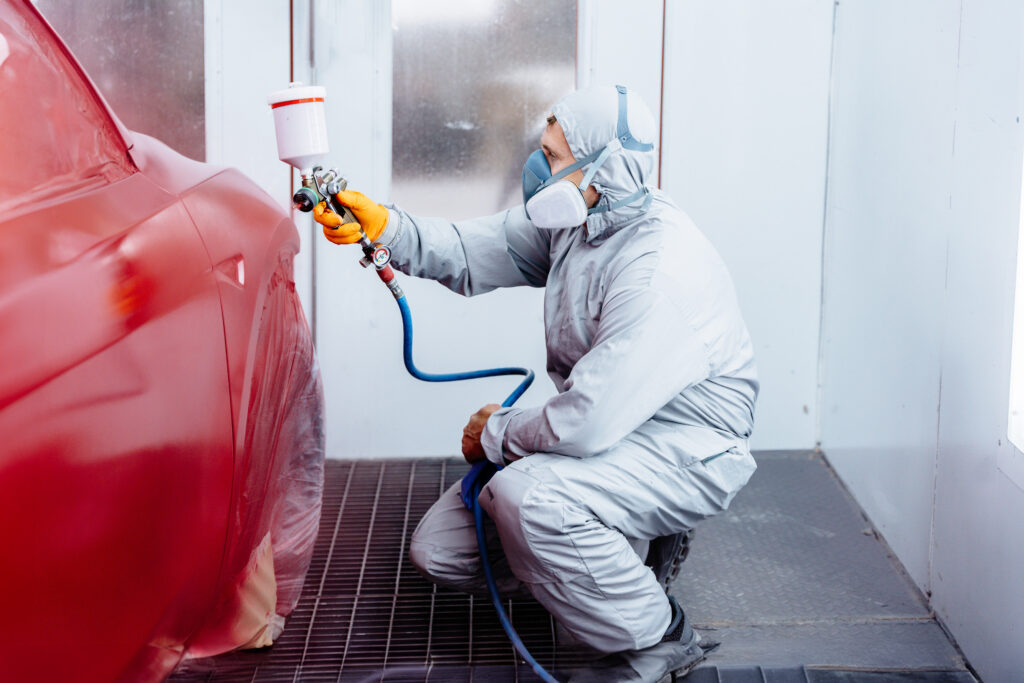 Peintre professionnel en combinaison et masque respiratoire, appliquant de la peinture rouge sur une voiture avec un pistolet.