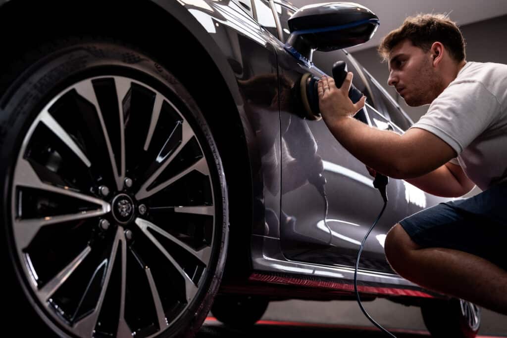 Homme poli méticuleusement une voiture grise foncée avec une machine. La carrosserie brillante reflète les lumières. Jante design visible.