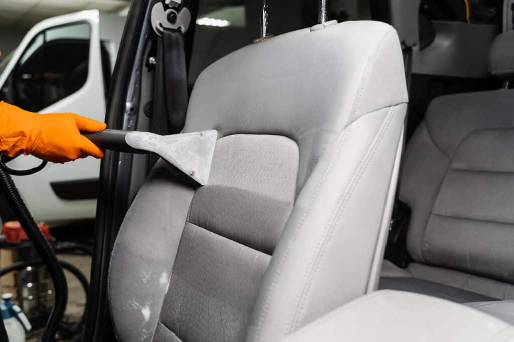 Main gantée de orange nettoyant un siège auto gris avec un extracteur, des zones mouillées apparentes. Nettoyage intérieur professionnel.