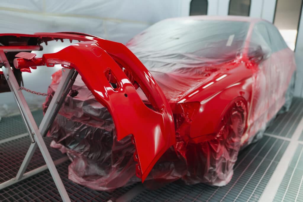 Voiture rouge et pare-chocs fraîchement peints en rouge vif, séchant dans une cabine de peinture automobile. La carrosserie est protégée par une bâche plastique.