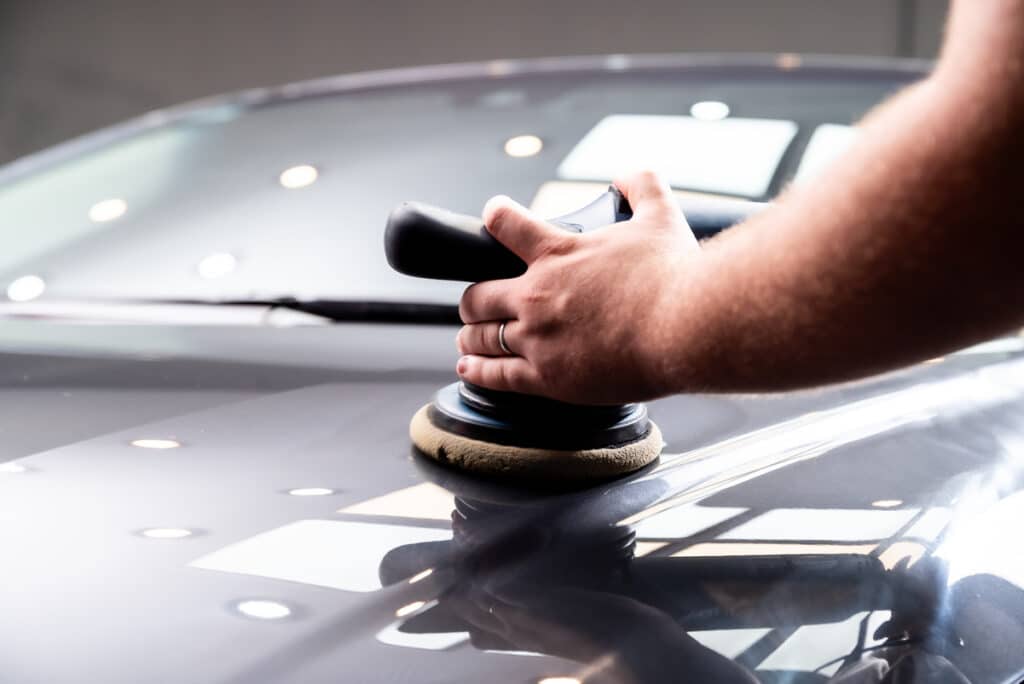 Main d'un homme polissant le capot brillant d'une voiture noire avec une polisseuse orbitale, reflétant des lumières vives.