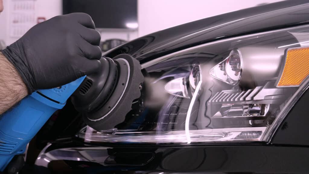Main d'un professionnel gantée polissant le phare et le capot noirs d'une voiture avec une lustreuse bleue. Surface brillante reflétant l'atelier.
