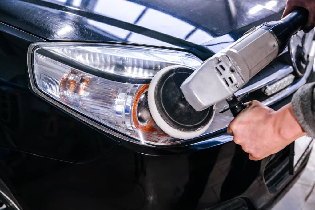Mains polissant un phare de voiture noire avec une machine. La carrosserie présente des reflets et des micro-rayures.