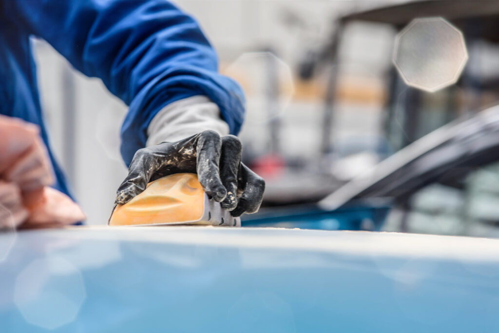Main gantée ponçant une carrosserie de voiture bleue avec un bloc orange. Poussière blanche visible, préparation automobile.
