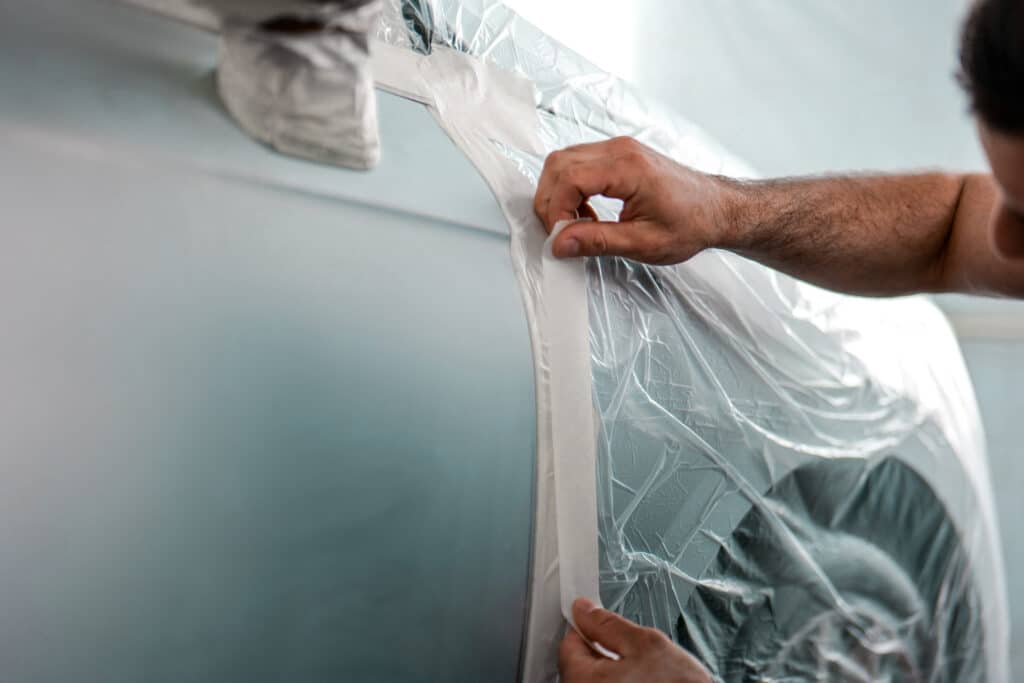 Mains appliquant du ruban de masquage sur une bâche plastique transparente recouvrant la vitre d'une voiture gris clair avant peinture.