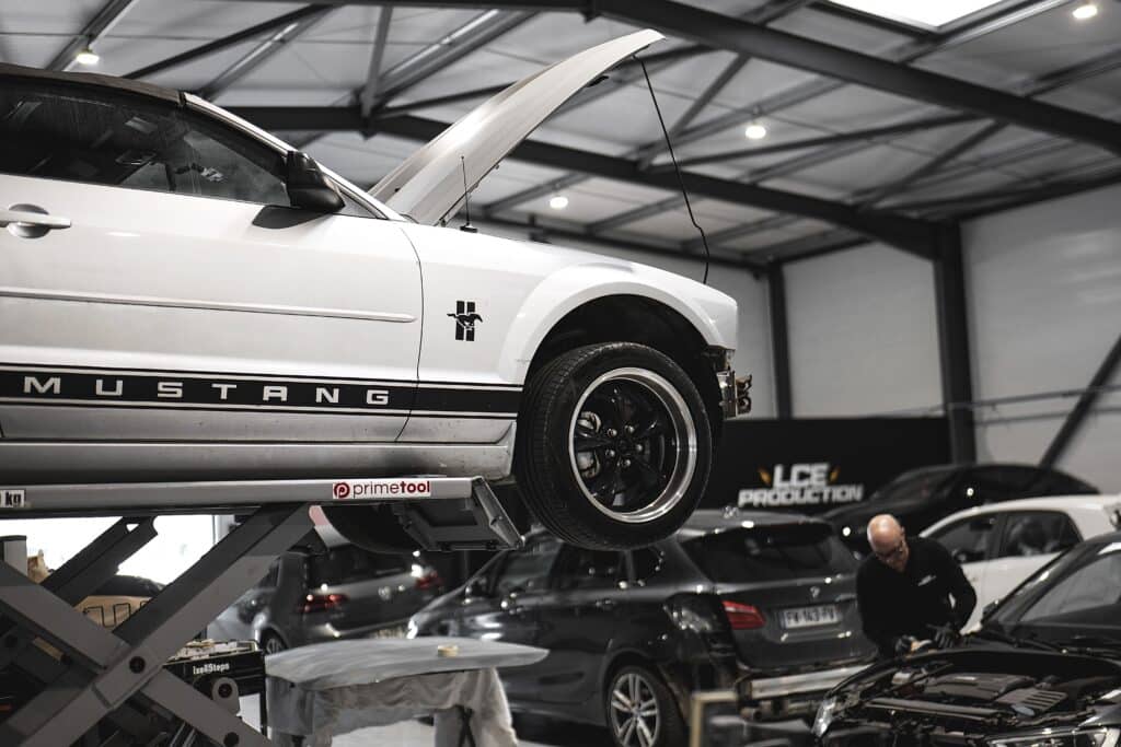 Mustang blanche sur pont élévateur Primetool, capot ouvert. Mécanicien travaillant sur moteur. Garage LCE Production.