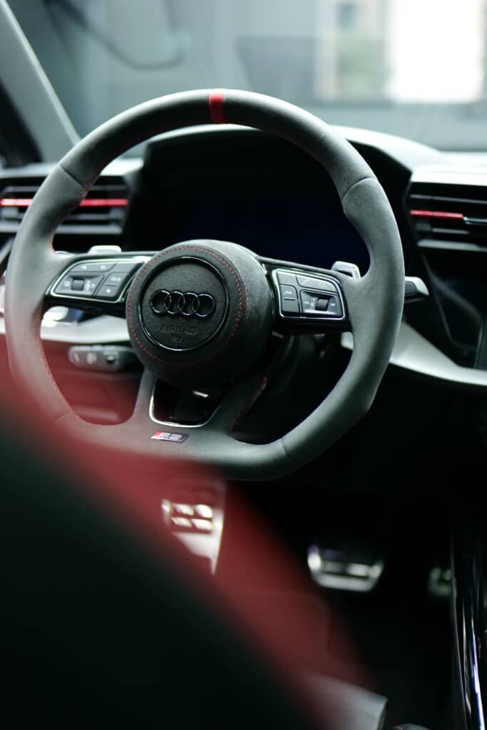 Volant sport Audi RS en Alcantara noir avec surpiqûres rouges, bande rouge à 12h et logo Audi. Commandes multifonctions et "AIRBAG" visibles.